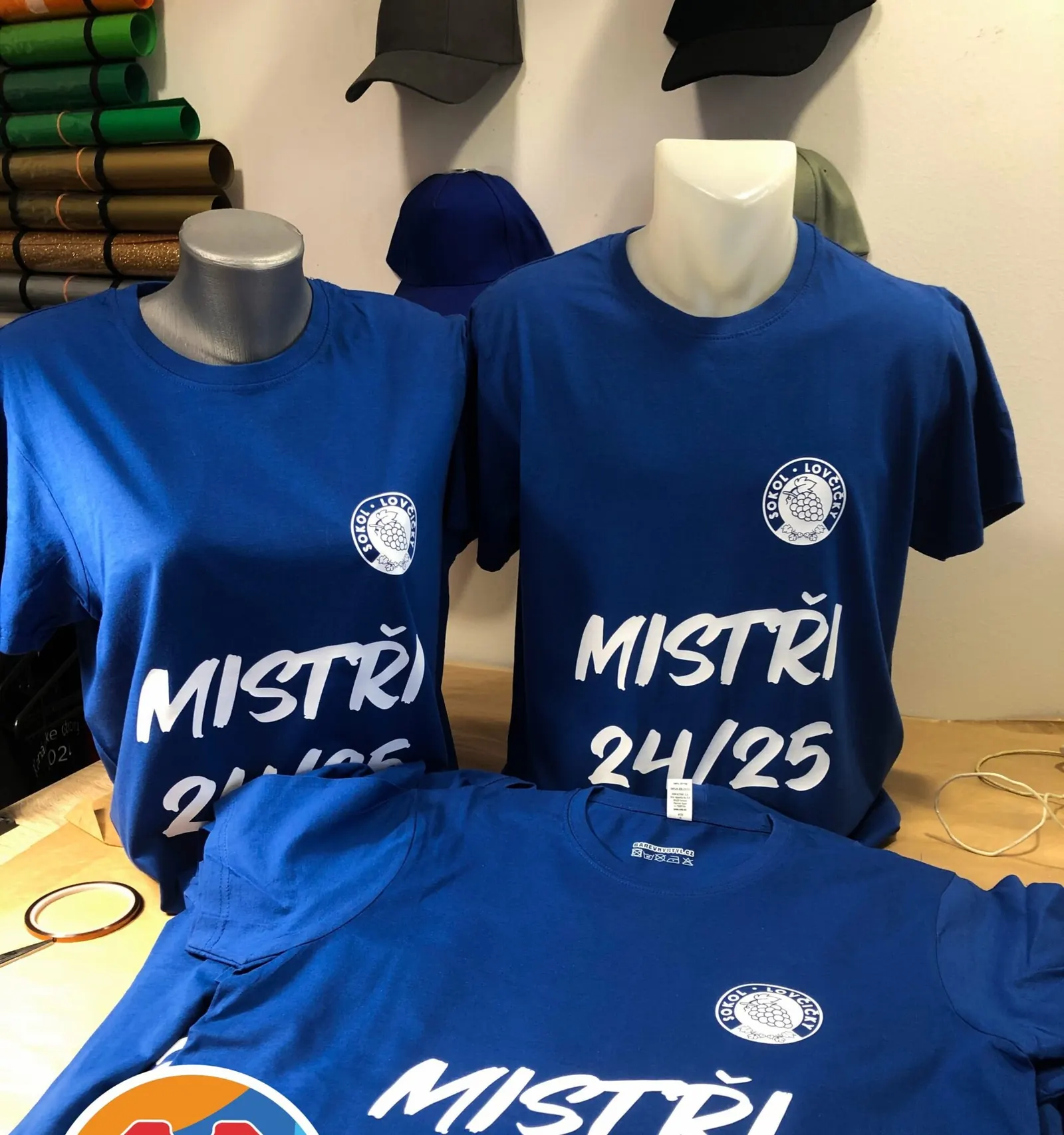 Modrá trička s bílým nápisem Mistři 24/25 na figurínách
